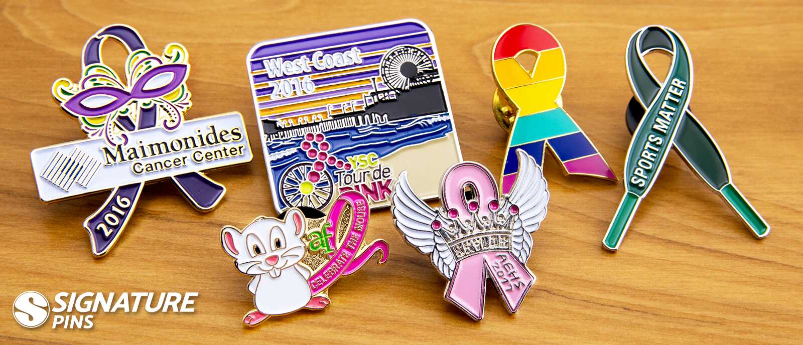 Custom awareness ribbon pins including Maimonides Cancer Center purple ribbon, Tour de Pink cycling pin, rainbow awareness ribbon, teal Sports Matter ribbon, and pink ribbon with wings and crown