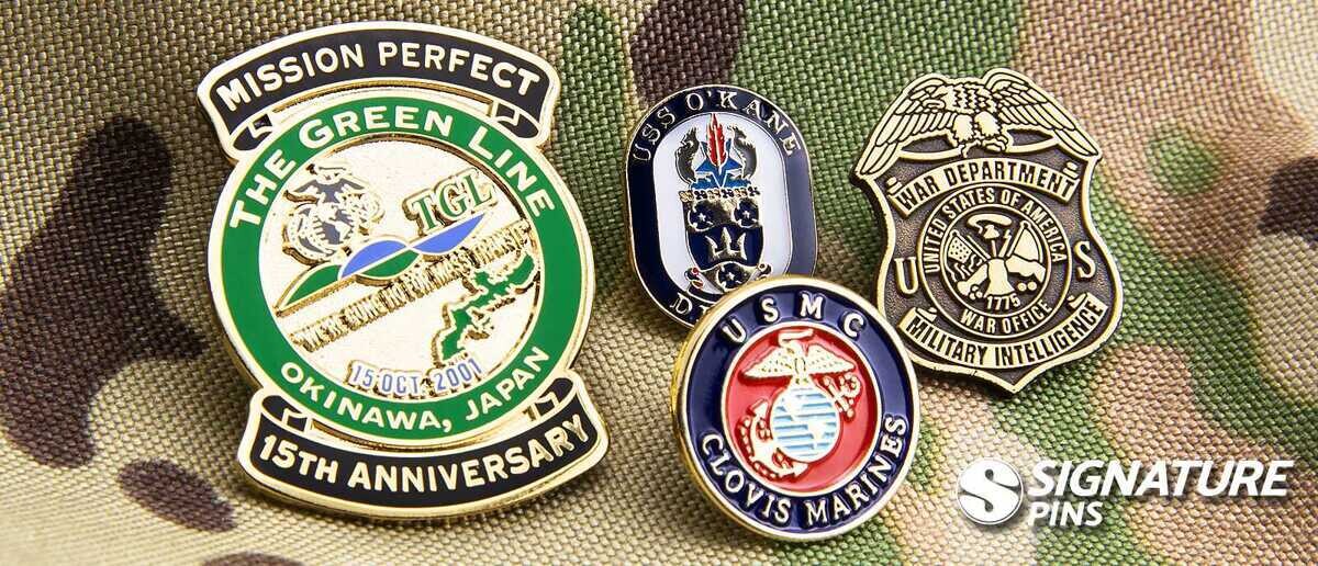 Collection of military pins on camouflage background including The Green Line 15th Anniversary Okinawa Japan pin, USS O'Kane DDG-77 pin, War Department Military Intelligence badge, and USMC Clovis Marines pin